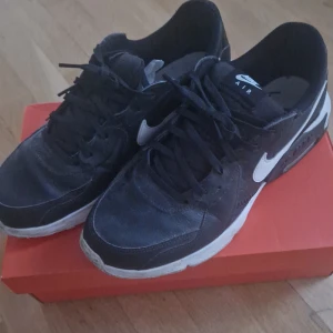 Svarta Nike Air Max sneakers - Säljer ett par svarta Nike Air Max sneakers med vit sula och klassisk swoosh på sidan. Skorna har snörning och en rund tå, samt synlig Air-dämpning i sulan. Materialet är en mix av syntet och skinn, vilket ger en sportig och clean look. Perfekta för dig som gillar streetstyle.