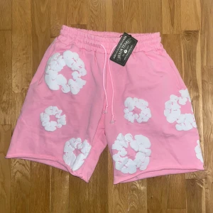 Rosa shorts från Denim Tears  - Säljer ett par rosa shorts från Denim Tears med vita blommiga tryck över hela. De har elastisk midja med snörning och är gjorda i mjuk bomull. Perfekt för chill dagar och sommarhäng. Snygg loose passform och unik design som sticker ut.