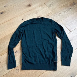 Mörkgrön merinotröja från BOSS - Står att storleken är L men är s .Slim fit mörkgrön stickad tröja från BOSS i extrafin merinoull. Tröjan har rund hals, långa ärmar och en liten metallbricka med BOSS-logga vid nederkanten. Perfekt för dig som gillar stilrena och sköna plagg med premiumkänsla.