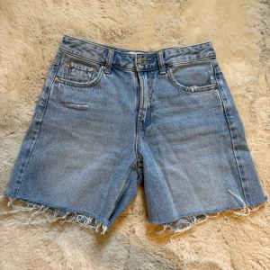 Blå jeansshorts från Stradivarius - Snygga blå jeansshorts från Stradivarius med medel midja och fransiga benslut. Klassisk femficksmodell i denim med knappgylf och dragkedja.