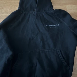 Svart Essentials Fear of God hoodie - Svart hoodie från Essentials Fear of God med tryck på bröstet och ärmen. Klassisk oversized passform, stor huva och magficka. Mjuk bomullskvalitet som är perfekt för chill dagar. Snygg streetwear-vibe och diskret branding på huvan.