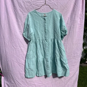 Ljusblå kortklänning från Rhythm - Supersöt ljusblå kortklänning från Rhythm med puffärmar och knappar framtill. Klänningen har en avslappnad passform, volangdetaljer och är gjord i en skön blandning av bomull och ramie. Perfekt för varma dagar när du vill känna dig både chill och stylish.