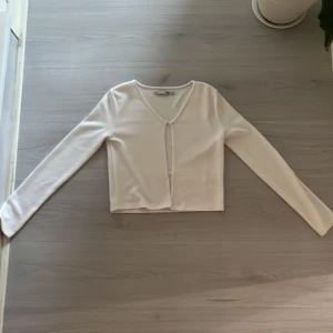 Vit ribbad kofta från 157 - Säljer en vit ribbad kofta från 157 med lång ärm och croppad passform. Koftan har v-ringad hals och en liten knäppning framtill. Perfekt att styla med jeans eller kjol för en clean look.