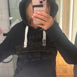 Svart Superdry hoodie med glitterlogga - Svart hoodie från Superdry med stor glittrig logga i guld och svart på bröstet. Tröjan har vit snörning i huvan och klassisk Superdry-patch på snöret. Skitsnygg!! Priset går att diskuteras 