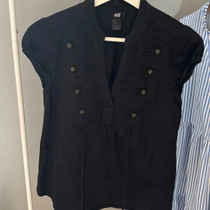 Svart blus med knappar från H&M - Svart blus från H&M. Den har korta ärmar, djup v-ringning och dekorativa knappar framtill. Materialet är mjukt och lätt, perfekt för dig som gillar stilrena detaljer och vill ha en unik topp i garderoben.