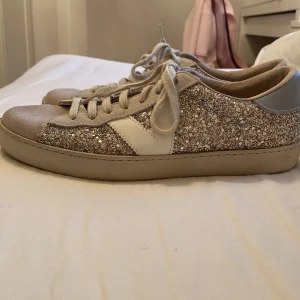 Glittriga beige sneakers med vit detalj - Säljer ett par snygga sneakers från Victoria med glittriga paneler i beige och silver, vit V-detalj på sidan och ljusgrå mocka vid tån. Skorna har rund tå, platt sula och klassisk snörning. Perfekta för dig som gillar att sticka ut med lite extra glitter.