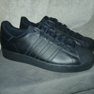 Svarta Adidas Superstar sneakers - Säljer ett par svarta Adidas Superstar sneakers i storlek 44. Skorna har klassisk snörning, rund tå och är tillverkade i skinn med matchande svarta detaljer och sula. Ikoniska tre ränder på sidan och Adidas-logga på hälen och plösen. Perfekt för dig som gillar stilrena och tidlösa sneakers.