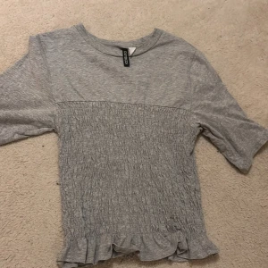 Grå ribbad topp med volang från H&M Divided - Supersöt grå topp från H&M Divided i storlek S. Toppen har ribbad struktur över bröstet och midjan, korta ärmar och en volangkant längst ner som ger en extra touch. Perfekt för dig som gillar enkel men ändå unik stil.Säljer den då den inte kommer till användning längre! Men super fräsch tröja!Pris kan diskuteras. Använt den några gånger, men ser nästan helt ny ut