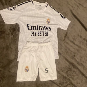 Real Madrid Bellingham #5 fotbollströja - Säljer en vit Real Madrid fotbollströja med Bellingham och nummer 5 på ryggen. Tröjan har korta ärmar, svarta detaljer och klubbmärke på bröstet. Materialet är lätt och andas, perfekt för match eller träning. Ingår matchande shorts med klubbmärke och nummer 5.