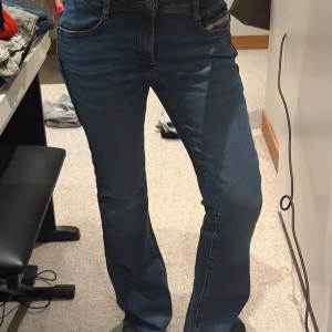 Diesel bootcut jeans blå - Snygga blå jeans från Diesel med klassisk bootcut-passform och fem fickor. Jeansen har en mörk tvätt med lätt slitning och detaljerad logotyp på fickan. Perfekt för dig som gillar en tidlös och cool stil. Materialet är mjukt jeans och midjan är normal.