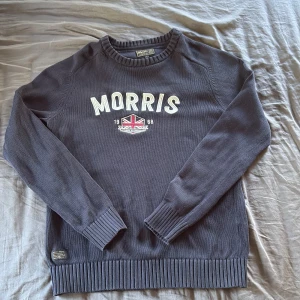 Mörkblå stickad tröja Morris S - Snygg mörkblå stickad tröja från Morris i storlek S. Tröjan har rund hals, långa ärmar och ett broderat Morris-tryck med brittisk flagga på bröstet. Ribbstickade muddar vid ärmslut och nederkant ger en schysst passform. Perfekt för dig som gillar klassisk stil.