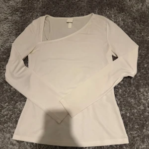 Vit tröja från H&M - Säljer en stilren vit topp från H&M i storlek S. Toppen har lång ärm och en snygg omlott-design med asymmetrisk halsringning. Materialet är mjukt och stretchigt, perfekt för dig som gillar minimalistisk och clean stil. Aldrig använt 