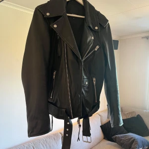 Klassisk Biker Läder Jacka från Rock’n Blue. - Säljer en svart skinnjacka från Rockandblue med cool bikerstil. Jackan har flera dragkedjor, bred krage, bälte i midjan och snygga silvriga detaljer. Insidan har en röd kant som ger extra edge. Perfekt för dig som vill ha en klassisk men ändå modern look.