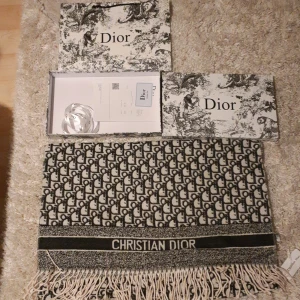 Svartvit halsduk från Christian Dior - Exklusiv halsduk från Christian Dior med ikoniskt monogrammönster i svart och vitt. Tjockt, mjukt material med fransar i kanterna och tydlig logga. Perfekt accessoar för att lyfta din outfit med lyxig vibe.