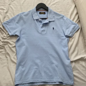 Ljusblå Polo Ralph Lauren t-shirt - Snygg ljusblå t-shirt från Polo Ralph Lauren med klassisk pikékrage och korta ärmar. Ikonisk broderad logga på bröstet och knappar vid halsen. Tillverkad i mjuk bomull som känns skön mot huden. Perfekt för dig som gillar stilren och sportig vibe.