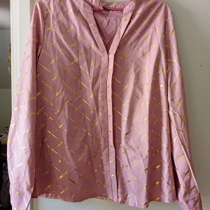 Rosa skjorta med guldmönster - Säljer en rosa långärmad skjorta med glittrigt guldmönster i rutigt snitt. Skjortan har v-ringad hals och är tillverkad i polyester.. Perfekt för dig som gillar att sticka ut med färg och glans.