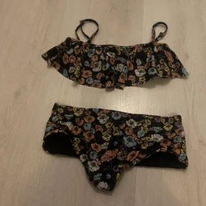 Blommig bikini med volang - Superfin bikini med blommigt mönster i orange, gul, blå, vit och lila. Toppen har volangdetalj och smala axelband. Bikinin har en klassisk form och spänne i ryggen. Perfekt för strandhäng och poolparty!