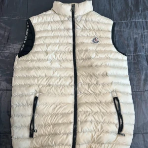 Beige dunväst från Moncler - Snygg beige dunväst från Moncler med svart dragkedja och två fickor framtill. Västen har hög krage och klassisk Moncler-logga på bröstet. Quiltad design och sportig look, perfekt att slänga över en hoodie eller tröja.