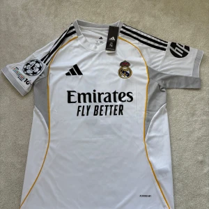 Real Madrid vit matchtröja Adidas S - Säljer en officiell Real Madrid matchtröja från Adidas i slim fit. Tröjan är vit med svarta och gula detaljer, broderat klubbmärke på bröstet och Champions League-patch på ärmen. Materialet är lätt och ventilerande Aeroready-polyester. Perfekt för dig som älskar fotboll och Real Madrid.