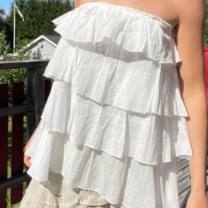 Vit volangig bandeau tubtopp - Supersöt vit bandeau tubtopp med flera lager volanger som ger en flowy och luftig känsla. Toppen är axelbandslös och har en lätt rynkad kant upptill för att sitta på plats. Perfekt för soliga dagar och sommarhäng. Står storlek 156/164 men skulle snarare säga xs-s! Går även att ha som kjol, likt att jag även säljer kjolen som går att ha som topp; två outfits i en!
