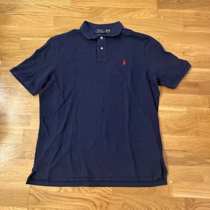 Mörkblå Polo Ralph Lauren t-shirt - Snygg mörkblå t-shirt från Polo Ralph Lauren med klassisk krage och två knappar framtill. Den har korta ärmar och det ikoniska röda Polo-logot broderat på bröstet. Perfekt för dig som gillar stilren och tidlös design.