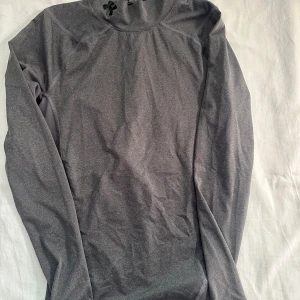 Grå långärmad träningströja Under Armour - Säljer en grå långärmad träningströja från Under Armour i storlek S. Tröjan är i kompressionsmodell med HeatGear-material som andas och transporterar bort fukt. Perfekt för gymmet eller löpning, med diskret logga på bröstet och ryggen.