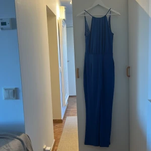 Lång blå jumpsuit med spets från Bik Bok - Mellanblå lång jumpsuit från Bik Bok i storlek XS.   Halterneck framtill som går ut till axelband. Djup och öppen i ryggen med fina spetsdetaljer och ärmlös design. Materialet är mjukt och lätt. Dragkedja i ryggen. Lite tecken på användning i form av noppor, se bild.
