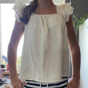 Beige blus med volangärmar - Säljer en söt beige blus med breda axelband och volangärmar med broderade detaljer. Blusen har en luftig passform och är gjord i ett lätt bomullstyg, perfekt för varma dagar. Volangerna ger en extra touch av stil och gör toppen riktigt charmig.