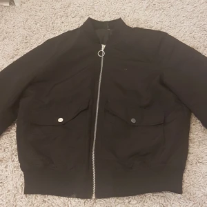 Svart bomberjacka med dragkedja - Svart bomberjacka med klassisk rund krage och silverfärgad dragkedja framtill. Jackan har två stora fickor med lock och tryckknappar, samt ribbade muddar vid ärmslut och nederkant. Perfekt för dig som gillar stilren och enkel streetwear.