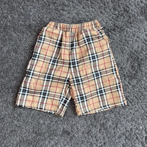 Beige rutiga shorts Burberry-style - Snygga beige shorts med klassiskt rutigt Burberry-mönster i svart, vitt och rött. De har elastisk midja för skön passform och är gjorda i mjuk bomull. Perfekta för dig som gillar ikoniska mönster och vill sticka ut med en trendig look.