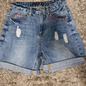 Blå jeansshorts med broderi Black Story - Snygga blå jeansshorts från Black Story med hög midja och coola slitningar framtill. Shortsens kanter är uppvikta och har rosa broderade detaljer både fram och bak. Klassisk femficksmodell med knapp och dragkedja. Jag har broderat blommorna själv. 