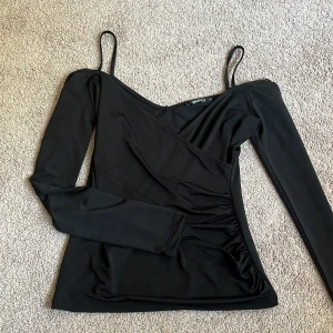 Svart omlottopp från Gina Tricot - Svart omlottopp från Gina Tricot i storlek XS. Toppen har långa ärmar och smala axelband, vilket ger en cool asymmetrisk look. Tillverkad i mjukt och stretchigt syntetmaterial som sitter snyggt på kroppen.