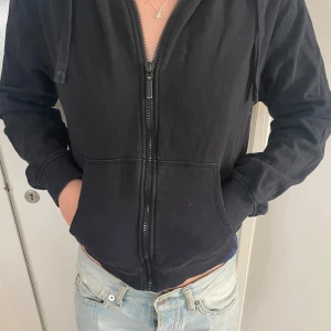 Svart hoodie med dragkedja - Svart hoodie med huva och hel dragkedja framtill. Tröjan har långa ärmar, ribbade muddar och fickor på sidorna. Materialet är mjuk bomull, perfekt för chill dagar. Enkel och stilren design som funkar till jeans eller joggers.