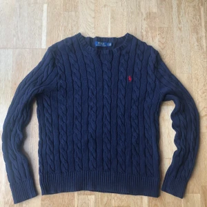 Marinblå kabelstickad tröja Polo - Kabelstickad marinblå tröja från Polo Ralph Lauren med klassisk rund hals och röd broderad logga på bröstet. Tröjan har långa ärmar och ribbade muddar vid ärmslut och nederkant. Har du någon fråga eller liknande är det bara att ställa.