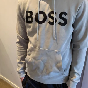 Boss hoodie  - Säljer en beige hoodie från BOSS i storlek S. Tröjan har svart BOSS-tryck på bröstet, huva med snörning och en stor magficka. Mjuk bomullskänsla och ribbade muddar vid ärmslut och nederkant. Perfekt för chill dagar och streetwear. Modellen är 180cm och väger 73kg