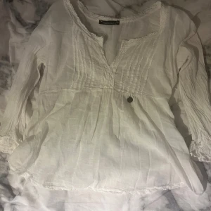 Vit blus med spets och v-ringning - Supersöt vit blus från Made in Italy i storlek S. Blusen har v-ringning med spetsdetaljer, smockade långa ärmar och fina veck framtill. Materialet är lätt och luftigt linne, perfekt för en chill och fräsch look. Liten medaljong-detalj vid midjan för extra stil.