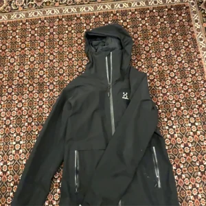 Svart vindjacka från Haglöfs med huva - Svart vindjacka från Haglöfs med snygg minimalistisk design, hel dragkedja och två praktiska fickor med dragkedja. Jackan har en justerbar huva och diskret logga på bröstet. Perfekt för dig som gillar outdoor och vill ha en stilren look.