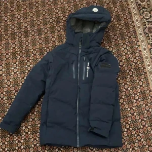 Mörkblå Sail Racing dunjacka med huva - Säljer en mörkblå dunjacka från Sail Racing med huva och snygga detaljer som Sail Antarctica Expedition-märke på ärmen. Jackan har flera praktiska fickor med dragkedja och justerbara muddar. Perfekt för kalla dagar och har en clean, sportig look.