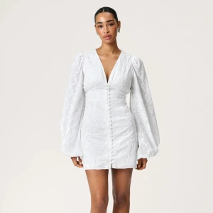 Adoore klänning  - Supersöt vit kort klänning med djup v-ringning och långa puffärmar. Klänningen har broderade blommor, knappar framtill och markerad midja som ger en snygg siluett. Perfekt för student eller sommarens alla tillfällen!