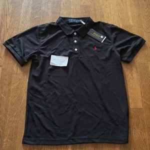 Svart Polo Ralph Lauren t-shirt S - Svart t-shirt från Polo Ralph Lauren med klassisk pikékrage och tre vita knappar framtill. Röd broderad logga på bröstet ger en clean och stilren vibe. Materialet är mjuk bomull som känns skönt mot huden. Perfekt för dig som gillar enkel och snygg design.
