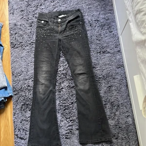 Svarta bootcut jeans med nitar från H&M - Snygga svarta jeans från H&M med bootcut och låg midja. Framtill är de dekorerade med små silvriga nitar som ger en cool vibe. Jeansen har klassisk femficksdesign, dragkedja och knapp, och är gjorda i stretchigt bomullsmaterial för skön passform. Väl andvända. Storlek 164 cm.