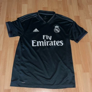 Svart Real Madrid fotbollströja Adidas - Snygg svart Real Madrid fotbollströja från Adidas med korta ärmar och klassiska ränder på axlarna. Tryck med Fly Emirates och klubbmärke på bröstet. Tillverkad i lätt och ventilerande polyester, perfekt för träning eller match.