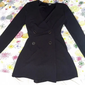 Svart omlottklänning med knappar - Snygg svart kortklänning med omlott-design och markerad midja. Klänningen har långa ärmar och fyra dekorativa knappar framtill som ger en cool, dressad look. Perfekt för dig som gillar stilrena och trendiga plagg med lite edge.