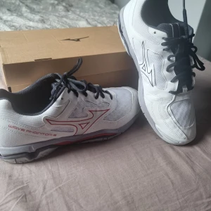 Mizuno Wave Phantom 3 sneakers vita - Snygga Mizuno Wave Phantom 3 sneakers i vitt med svarta och röda detaljer. Skorna har svart snörning, meshpaneler för andning och en slitstark sula med rött inslag. Perfekta för träning eller sport. Klassisk Mizuno-logga på sidan och häl. Lätt använda men inga skador