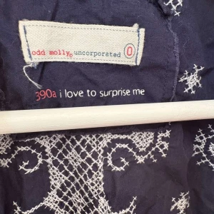 Odd Molly mönstrad kortklänning - Supersnygg kortklänning från Odd Molly med marinblå botten och vita broderade mönster. Klänningen har beige detaljer vid midjan och v-ringad hals, samt ett satinband som knyts i ryggen. Långärmad och med coola broderier som ger en unik vibe.