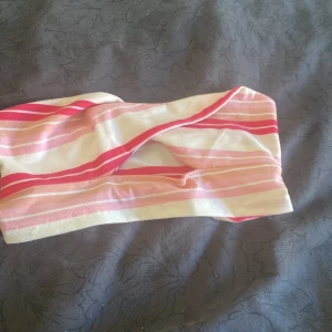 Randig bandeau tubtopp i rosa och vit - Säljer en söt bandeau tubtopp med randigt mönster i olika nyanser av rosa och vitt. Toppen är axelbandslös och har en twistad detalj framtill som ger en cool vibe. Perfekt för varma dagar och att styla med jeans eller shorts.