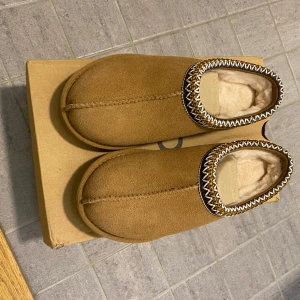 Bruna tofflor från UGG - Säljer ett par bruna tofflor från UGG i mocka med mjukt foder. De har en dekorativ kant med mönster och en bekväm sula. Perfekta för att hålla fötterna varma och sköna.