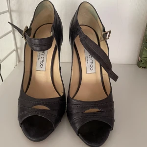 Mörkblå/guld  klackskor från Jimmy Choo - Snygga svarta klackskor från Jimmy Choo med öppen tå och justerbart spänne runt vristen. Skorna har en elegant design med hög klack och är perfekta för speciella tillfällen. Köpta i New York för 5200kr