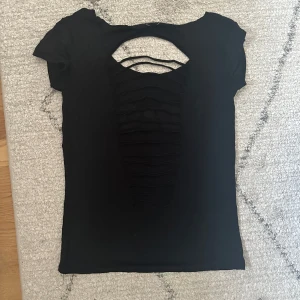 Svart kortärmad topp från D&G - Snygg svart topp från D&G med korta ärmar och ett coolt utskuret mönster på ryggen. Framsidan pryds av ett stort D&G-tryck i vitt. Perfekt för en trendig look! Står stl L men skulle säga xs/s