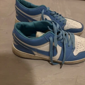 Jordan 1 - Snygga blå och vita Nike sneakers med snörning. Perfekta för en avslappnad stil. Skorna har en klassisk design med perforerade detaljer på ovandelen och en bekväm sula.